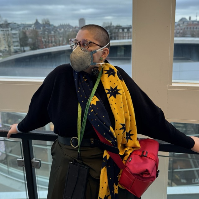 Anna, a white femme person, poses for a picture in the Van Gogh Museum in Amsterdam. They are leaning against a handrail installed in an access to stairs outside the galleries with their arms open, their body facing the camera, their face slightly turned to their right. Their hair is dark and short, almost completely shaved. They're wearing prescription glasses with tortoise frames, a grey N95 mask, a black turtleneck with a double face scarf with illustrations of expressive cartoonish stars in bright yellow and navy blue, a dark green skirt with a black belt. They're using a bright red purse across their body and an earring that imitates Van Gogh's Sunflowers. In the background, the cityscape of a cloudy Amsterdam makes a contrast against Anna's colorful accessories.